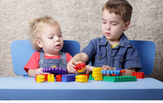 Zwei kleine Kinder, die an einem Tisch sitzen und darauf mit bunten Bausteinen spielen.