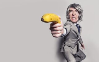 Ein Mann in einem grauen Anzug, der eine Banane wie eine Pistole ins Bild hält.