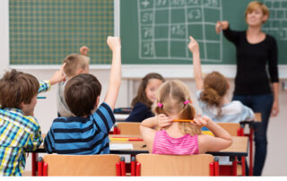 Kinder in der Schule, die sich melden. Im Hintergrund unscharf eine Lehrerin vor einer Tafel stehend.