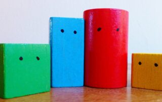 Vier bunte, nebeneinander aufgestellte Holzbausteine mit aufgemalten Augen.
