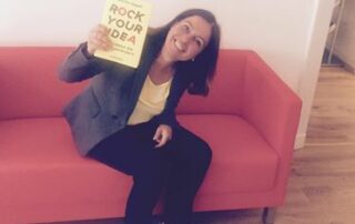Svenja Hofert, sitzend auf einem roten Sofa, hält lächelnd das Buch "Rock your idea" hoch.