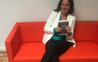 Svenja Hofert sitzt lächelnd auf einem roten Sofa und hält das Buch "Humble Consulting" in der Hand.