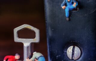 Eine Nahaufnahme eines Schlosses, dessen Schlüssel davor auf dem Boden steht. Im Schlüsselloch sitzt eine Figur, zwei weitere stehen am Schlüssel.