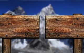Zwei Holzpfeiler, die genau in die gegengesetzte Richtung führen. Im Hintergrund unscharf Berge.
