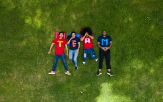 Vier Teammitglieder mit farbigen T-shirt mit jeweils einem Buchstaben, die insgesamt das Wort Team ergeben, liegen auf einer Wiese.