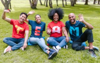 Vier Teammitglieder mit farbigen T-shirt mit jeweils einem Buchstaben, die insgesamt das Wort Team ergeben, sitzen auf einer Wiese und winken.