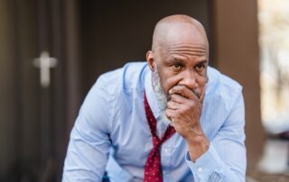 Businessman mit weißem Bart in Hemd und Krawatte sitzt nachdenklich.