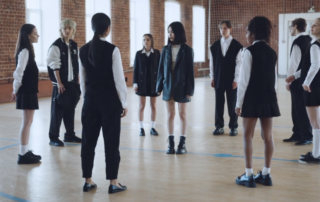 Gruppe junger Menschen in schuluniform-artigen Klamotten stehen in einer Sporthalle im Kreis. Ein Mädchen steht in der Mitte.