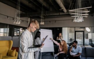 Ein Businessmeeting mit diskutierenden Teammitgliedern am Plipchart und einem Teammitglied am Laptop im Vordergrund.