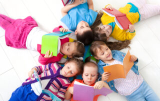 Sechs bunt gekleidete Kinder liegen im Kreis mit den köpfen in der Mitte und lesen.