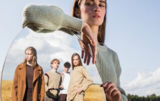 Eine junge Frau, die mit einem Spiegel posiert in dem andere Menschen zusehen sind. Ein Sinnbild für das psychologische Konzept von Übertragung und Gegenübertragung.